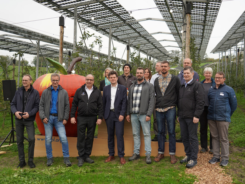 Ca 12 Menschen stehen vor der Agri-Photovoltaik-Anlage am FiBL. Sie stehen in zwei Reihen, die hintere Reihe steht auf einem Podest, auf dem auch ein mannshoher Apfel steht. Die Menschen tragen legere Hemden und Anzüge. Es handelt sich um viele Männer im besten Alter und einige Frauen. Die Menschen lächeln etwas schüchtern in die Kamera. Im Hintergrund sieht man gross die Solarpanels über der Niederstamm-Apfelplantage.